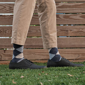 A relaxed standing pose on grass showing Blue Argyle dress socks paired with khaki pants and black leather derby shoes against a wooden fence backdrop. [lf]