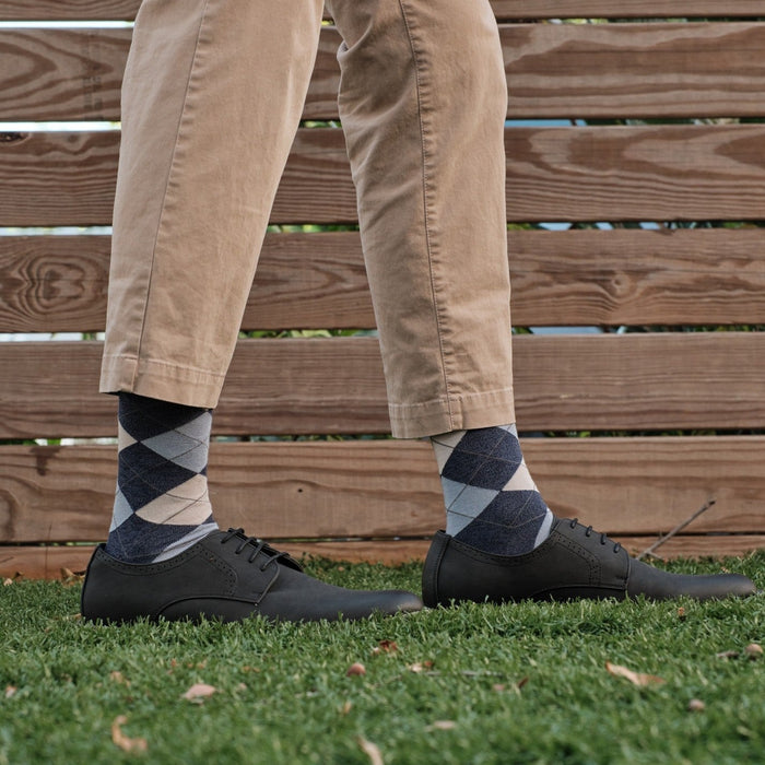 A relaxed standing pose on grass showing Blue Argyle dress socks paired with khaki pants and black leather derby shoes against a wooden fence backdrop. [lf]