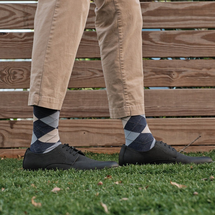 A relaxed standing pose on grass showing Blue Argyle dress socks paired with khaki pants and black leather derby shoes against a wooden fence backdrop. [lf]