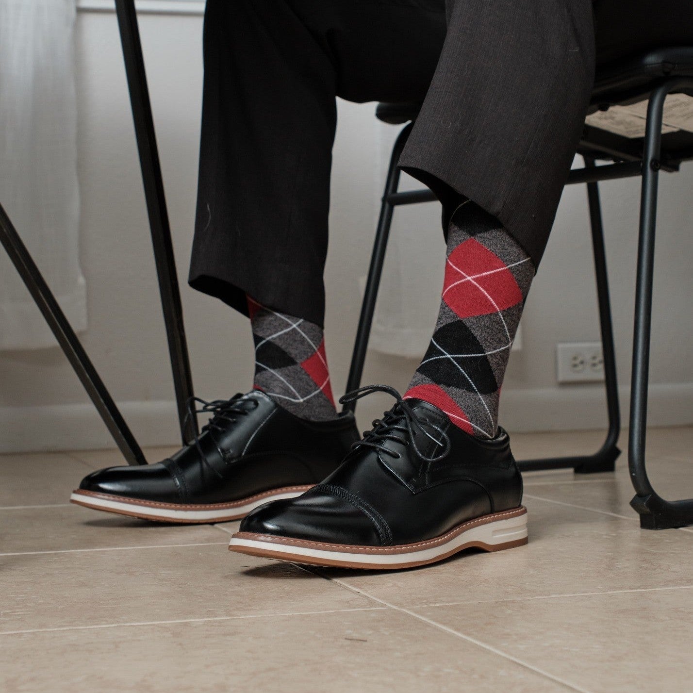 Charcoal Heather Argyle dress socks with bold red diamonds shown casually crossed at a desk, paired with grey dress slacks and black leather dress shoes. [lf]