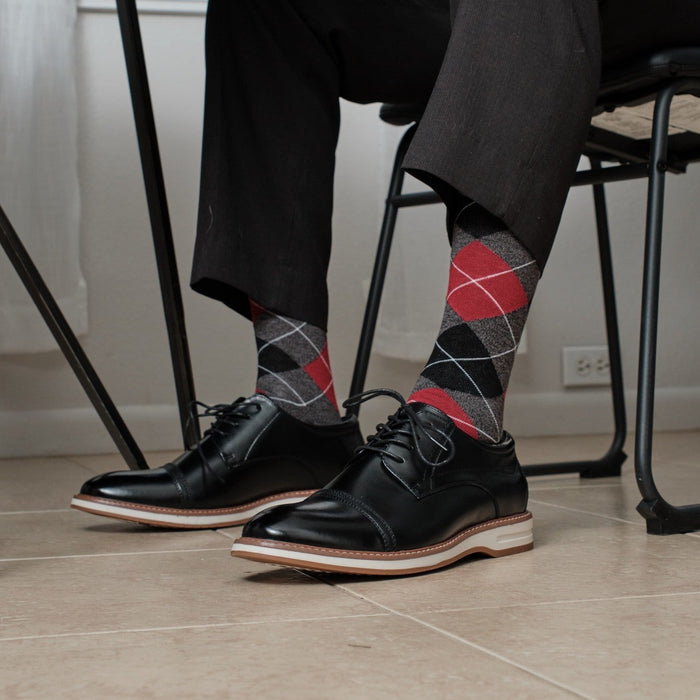 Charcoal Heather Argyle dress socks with bold red diamonds shown casually crossed at a desk, paired with grey dress slacks and black leather dress shoes. [lf]