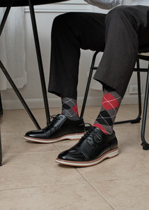 Close-up of Charcoal Heather Argyle dress socks with red and grey diamonds visible under a dining table, paired with grey trousers and black cap-toe oxfords. [lf]