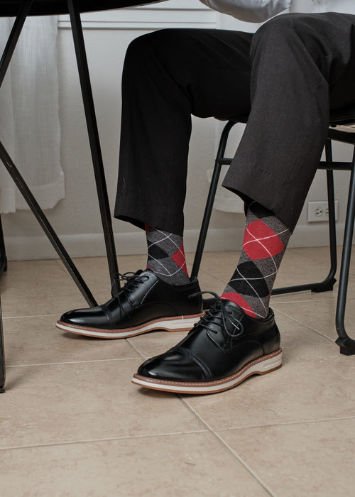 Close-up of Charcoal Heather Argyle dress socks with red and grey diamonds visible under a dining table, paired with grey trousers and black cap-toe oxfords. [lf]
