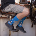 A man perched on a leather captain's chair steers a boat with his blue Shark Attack socks visible above black Vans sneakers, dressed in grey cargo shorts and a dark tee. [lf]