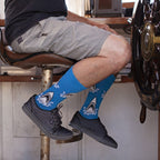 A man perched on a leather captain's chair steers a boat with his blue Shark Attack socks visible above black Vans sneakers, dressed in grey cargo shorts and a dark tee. [lf]