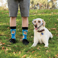 A man in plaid shirt and grey shorts stands on a grassy lawn beside a yellow Labrador wearing safety goggles, his Blue Science Lab socks featuring goggled lab dog prints visible above black sneakers. [lf]