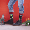 A person in cuffed jeans and dark sneakers stands against a bright red wall flanked by toy dinosaurs, wearing Black Santasaurus Rex socks patterned with festive T-rex prints, snowflakes, and Christmas trees. [lf]