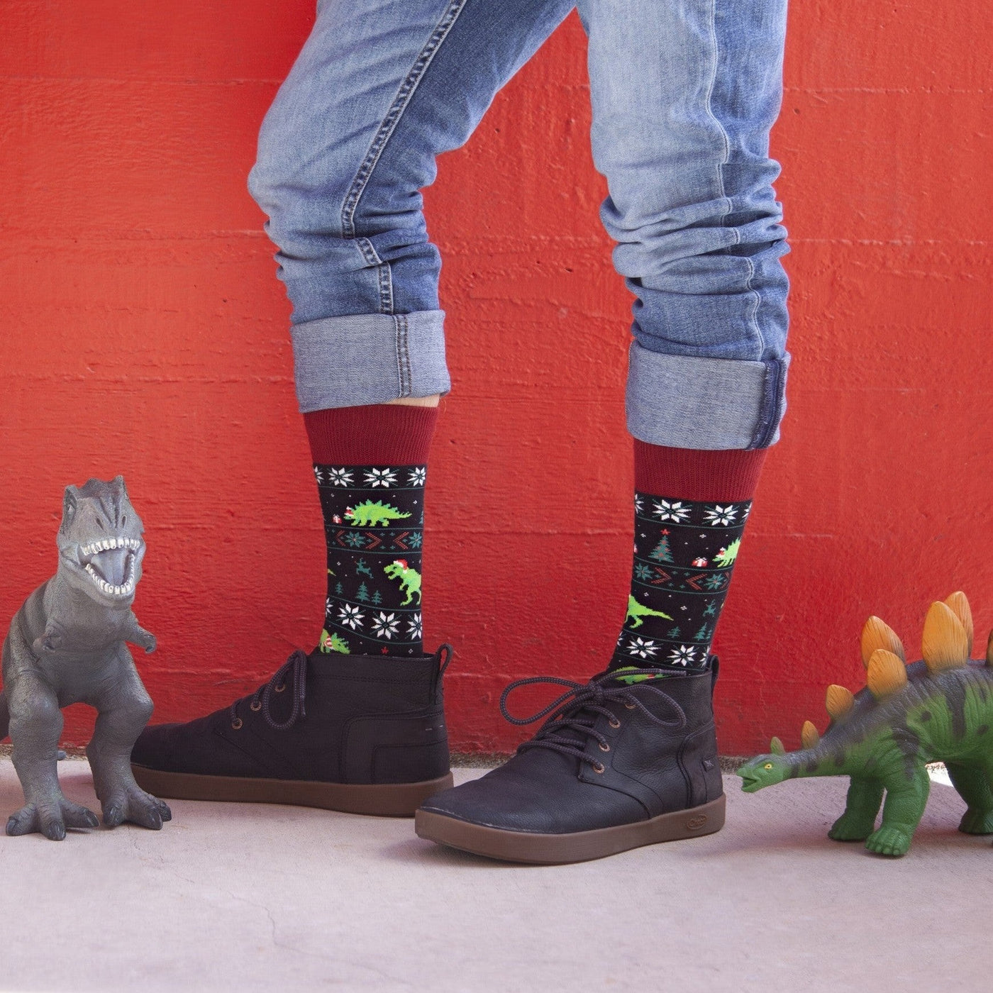 A person in cuffed jeans and dark sneakers stands against a bright red wall flanked by toy dinosaurs, wearing Black Santasaurus Rex socks patterned with festive T-rex prints, snowflakes, and Christmas trees. [lf]