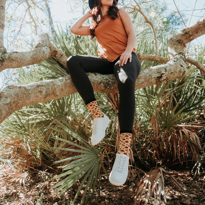 A woman in an orange tank top, black wide-brimmed hat, and black leggings perches on a curving tree branch over palm fronds, her white sneakers showing Ivory Leopard Print Bamboo socks with tan and black leopard spots. [lf]