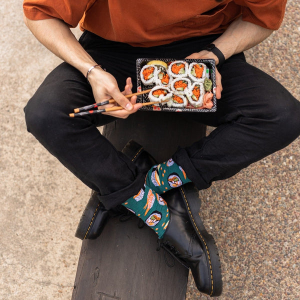 Overhead view of a man in an orange tee and black jeans holding chopsticks over a takeout tray of sushi rolls, his Teal Heather Sushi Come, Sushi Go Bamboo socks with sushi roll prints showing above black Doc Martens. [lf]