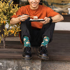 A smiling man sits on a porch step in an orange tee and black jeans, eating sushi with chopsticks beside a kumquat tree, his Teal Heather Sushi Come, Sushi Go Bamboo socks paired with black Doc Martens. [lf]