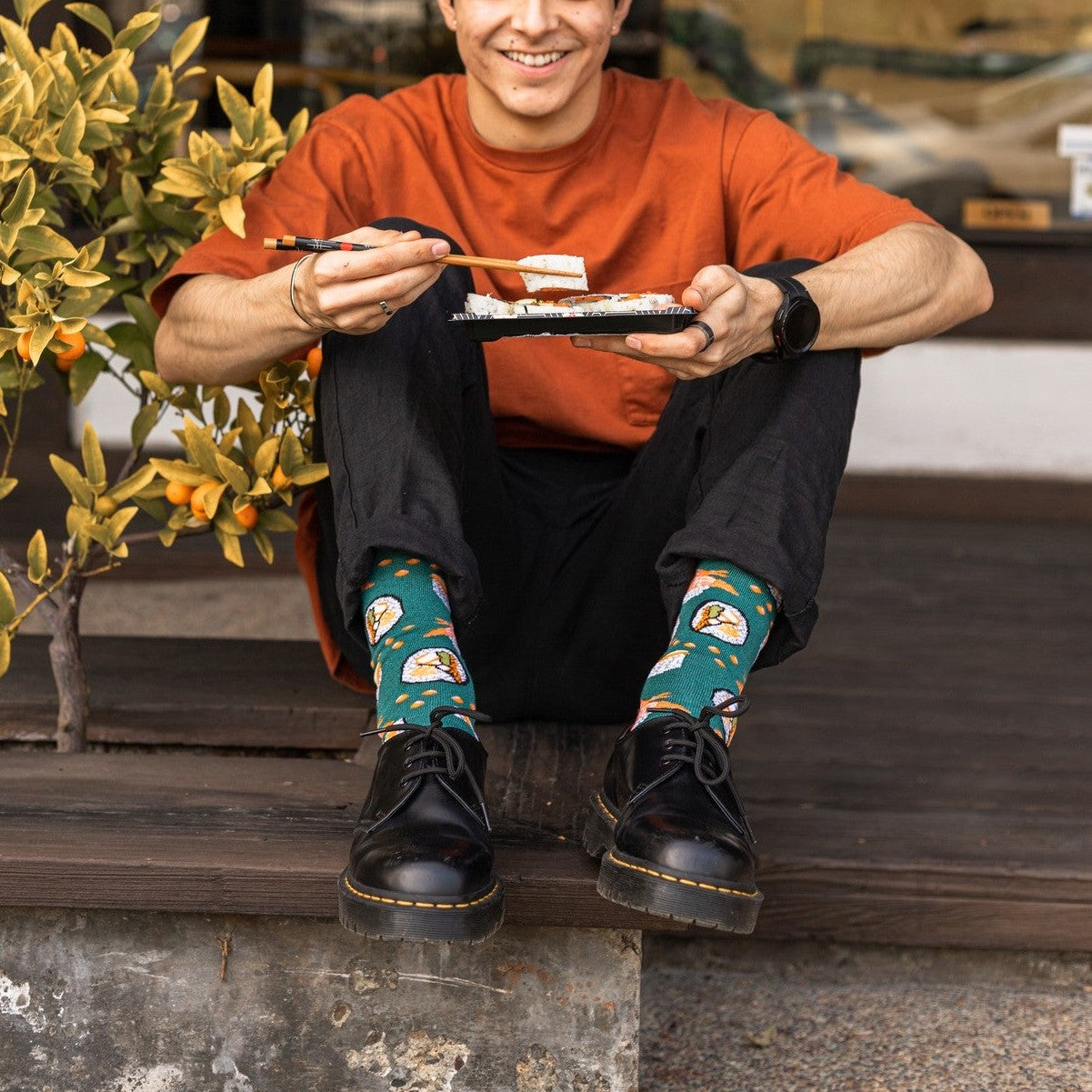 A smiling man sits on a porch step in an orange tee and black jeans, eating sushi with chopsticks beside a kumquat tree, his Teal Heather Sushi Come, Sushi Go Bamboo socks paired with black Doc Martens. [lf]