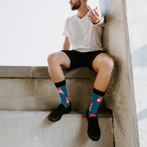 A man sits on a concrete parking-garage ledge in a white tee and black shorts, gesturing toward the camera with his Blue Can of Whoop Ass socks visible above black slip-on sneakers. [lf]