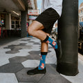 A man steps off a sidewalk on a New Orleans-style street, his Blue Can of Whoop Ass socks featuring red and white can prints peeking out of black slip-on sneakers paired with black shorts. [lf]