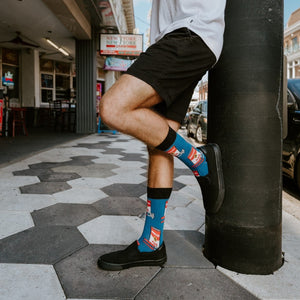 A man steps off a sidewalk on a New Orleans-style street, his Blue Can of Whoop Ass socks featuring red and white can prints peeking out of black slip-on sneakers paired with black shorts. [lf]