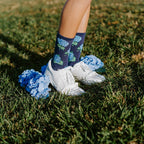 Close-up of bare legs in white sneakers on green grass, wearing Navy Hydrangeas socks with blue hydrangea bloom prints beside a real blue hydrangea blossom. [lf]