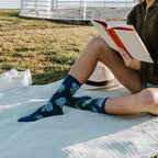 Curled up on a picnic quilt with a book, a woman in a brown sherpa and denim shorts shows off Navy Hydrangeas socks with blue hydrangea blooms on a sunny grass hillside. [lf]