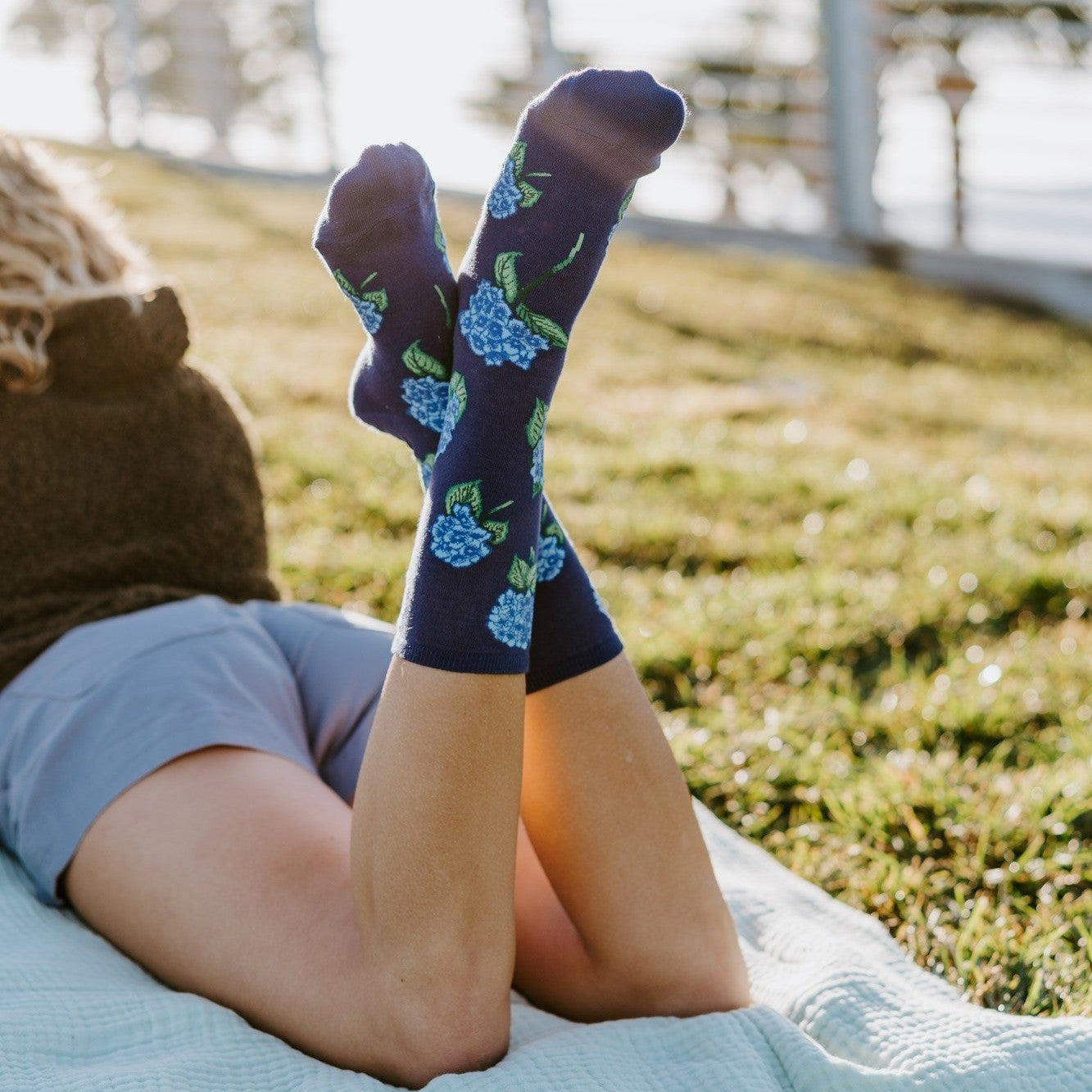 A close-up of crossed feet pointed skyward in Navy Hydrangeas socks with blue hydrangea prints, with the woman lounging on a quilt under a sunny coastal sky. [lf]