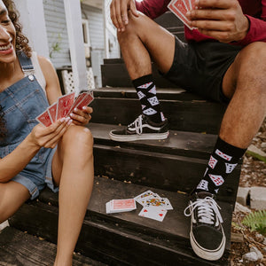 Close-up of two people playing cards on dark wooden porch steps, the man's Black Deal Me In Bamboo socks with card-suit prints visible above black Vans Old Skool sneakers. [lf]