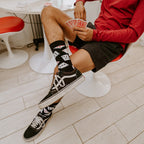Seated on a red tulip chair beside a snake plant, a man in a red long-sleeve and black shorts crosses his Black Deal Me In Bamboo socks with playing-card prints above black Vans sneakers. [lf]