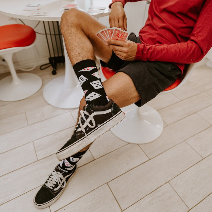 Seated on a red tulip chair beside a snake plant, a man in a red long-sleeve and black shorts crosses his Black Deal Me In Bamboo socks with playing-card prints above black Vans sneakers. [lf]