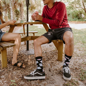 A young couple plays cards at a backyard picnic table, the man in a red shirt and Black Deal Me In Bamboo socks with card-suit prints crossing his legs in black Vans Old Skools. [lf]
