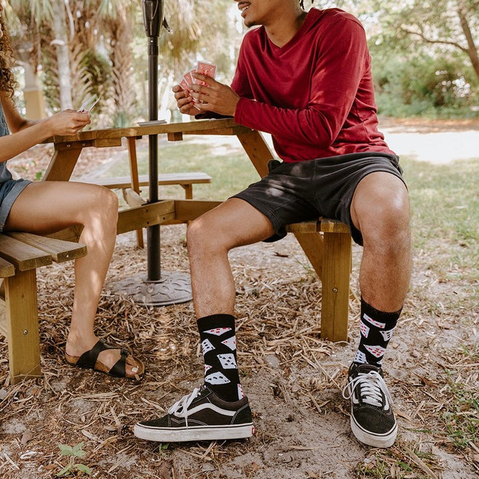 A young couple plays cards at a backyard picnic table, the man in a red shirt and Black Deal Me In Bamboo socks with card-suit prints crossing his legs in black Vans Old Skools. [lf]
