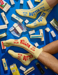 A person's legs and feet wearing pale yellow novelty socks printed with 'BUTTER' and 'NET WT. 4 OZ.