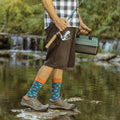 Vertical angle from chest down on a streamside fisherman gripping a spinning reel and green tackle case, his Just Fishin' socks in STEEL BLUE peeking from rolled shorts as he balances on wet rocks. [lf]