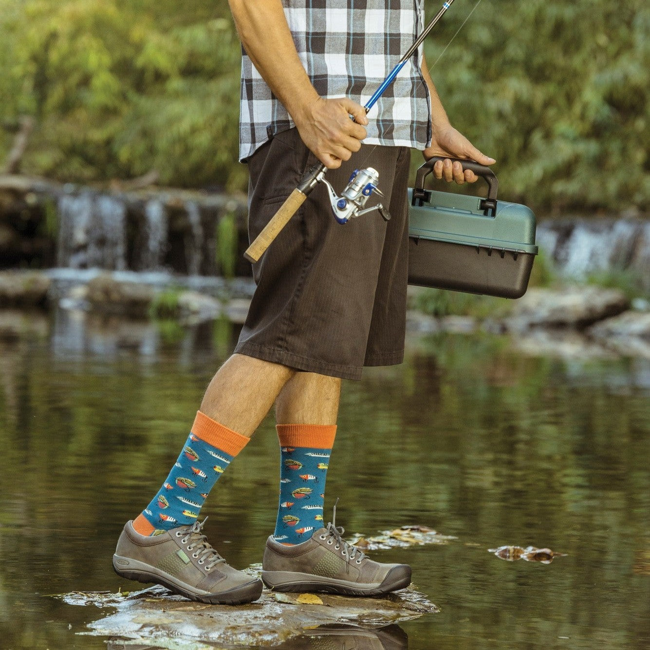 Vertical angle from chest down on a streamside fisherman gripping a spinning reel and green tackle case, his Just Fishin' socks in STEEL BLUE peeking from rolled shorts as he balances on wet rocks. [lf]