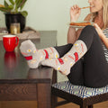 Vertical frame of her seated on a patterned cushion under a crowded photo gallery wall, crossed legs resting on the table to put Good Morning socks and their breakfast print at the heart of the shot. [lf]