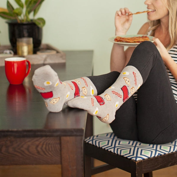 Vertical frame of her seated on a patterned cushion under a crowded photo gallery wall, crossed legs resting on the table to put Good Morning socks and their breakfast print at the heart of the shot. [lf]