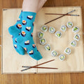 Overhead flat-lay of crossed feet on a light wooden tray next to a heart shaped from avocado sushi rolls and two pairs of chopsticks, wearing the Love At First Bite socks in turquoise printed with heart-shaped sushi rolls. [lf]