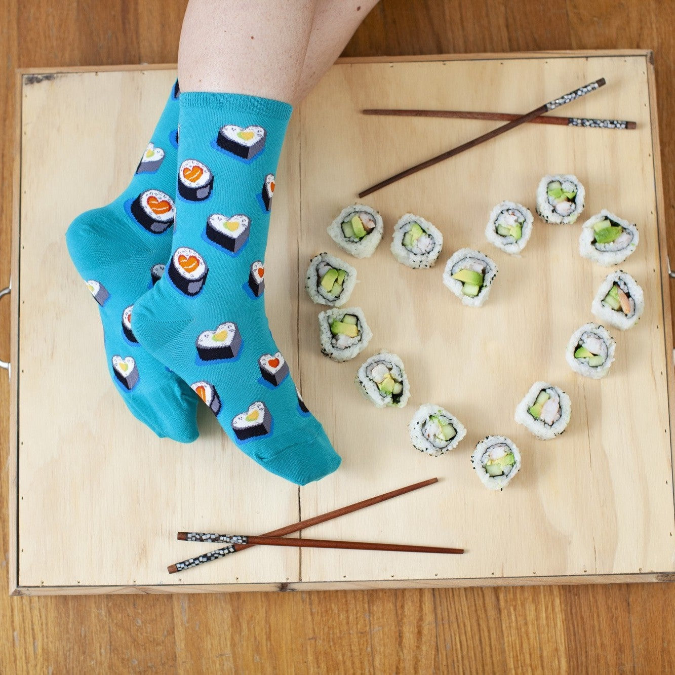 Overhead flat-lay of crossed feet on a light wooden tray next to a heart shaped from avocado sushi rolls and two pairs of chopsticks, wearing the Love At First Bite socks in turquoise printed with heart-shaped sushi rolls. [lf]