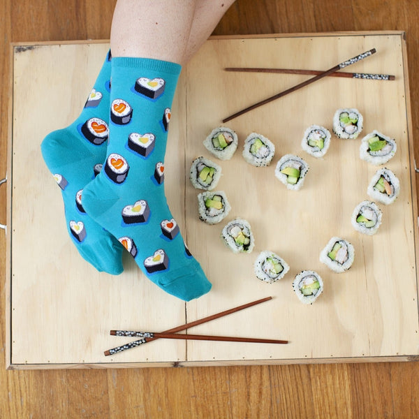 Overhead flat-lay of crossed feet on a light wooden tray next to a heart shaped from avocado sushi rolls and two pairs of chopsticks, wearing the Love At First Bite socks in turquoise printed with heart-shaped sushi rolls. [lf]