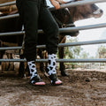A person stands at a livestock pen petting a brown cow while wearing MOOOO! in White socks with sandals. [lf]