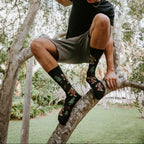 A young man perches on a tree branch, leaning down with one arm gripping a limb, wearing Bigfoot XL in BLACK socks with sneakers in a sunlit backyard. [lf]