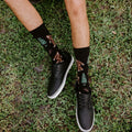 A top-down look at both feet standing on grass highlights the tree and sasquatch motifs of Bigfoot XL in BLACK socks paired with black sneakers. [lf]