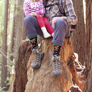 A father holds his daughter close on a sun-dappled stump surrounded by towering redwoods, their hiking boots topped with Big Foot Christmas socks in Black. [lf]