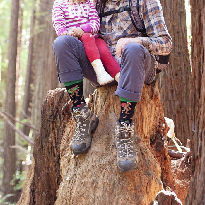 A father holds his daughter close on a sun-dappled stump surrounded by towering redwoods, their hiking boots topped with Big Foot Christmas socks in Black. [lf]