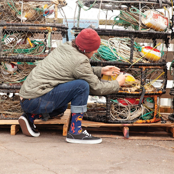 Wearer kneels at dockside crab traps tying gear, Seafood Platter socks peeking above black sneakers in profile view. [lf]
