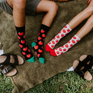 Two pairs of feet lined up on a picnic blanket showcase Apple Of My Eye socks in black with green toes and in white with red toes, with black and brown buckled sandals kicked off beside them. [lf]
