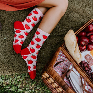 A woman's crossed legs in white Apple Of My Eye socks with bright red apple prints and red toes stretch out beside an open wicker picnic basket filled with apples, grapes, cheese and a baguette. [lf]
