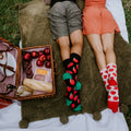 A couple lies side by side on their stomachs on a picnic blanket with feet kicked up, his black Apple Of My Eye socks with green toes next to her white pair with red apple prints and red toes. [lf]
