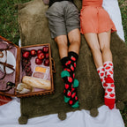 A couple lies side by side on their stomachs on a picnic blanket with feet kicked up, his black Apple Of My Eye socks with green toes next to her white pair with red apple prints and red toes. [lf]