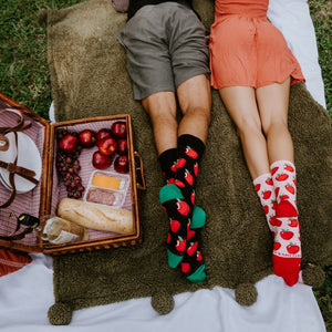 A couple lies side by side on their stomachs on a picnic blanket with feet kicked up, his black Apple Of My Eye socks with green toes next to her white pair with red apple prints and red toes. [lf]