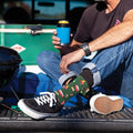 A man sits in the open tailgate of a pickup truck holding a koozie-wrapped can next to a green cooler and small black charcoal grill, wearing the Tailgaters Delight in Green Heather socks printed with hot dogs and burgers above black-and-white sneakers. [lf]
