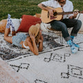 A young woman in a denim shirt and shorts lounges face-down on a patterned picnic rug wearing blue Blueberry Tie Dye Athletic socks, while a man with long hair sits in a chair playing acoustic guitar beside her in matching blue tie-dye socks. [lf]