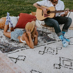 A young woman in a denim shirt and shorts lounges face-down on a patterned picnic rug wearing blue Blueberry Tie Dye Athletic socks, while a man with long hair sits in a chair playing acoustic guitar beside her in matching blue tie-dye socks. [lf]