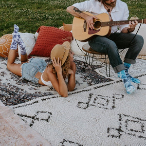 A young woman in a denim shirt and shorts lounges face-down on a patterned picnic rug wearing blue Blueberry Tie Dye Athletic socks, while a man with long hair sits in a chair playing acoustic guitar beside her in matching blue tie-dye socks. [lf]